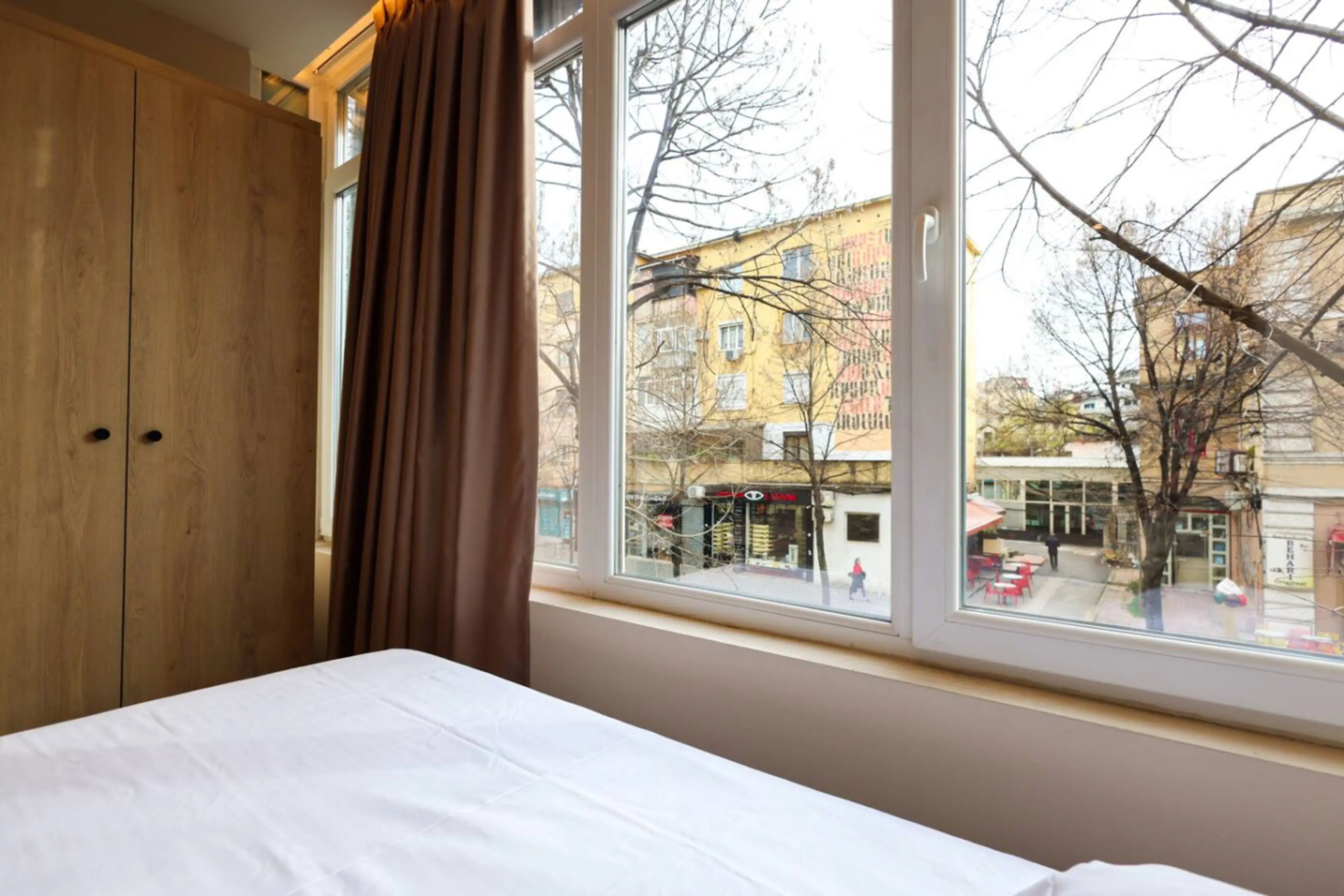 A bedroom with a bed, a wooden wardrobe, and a window with a view of the street.