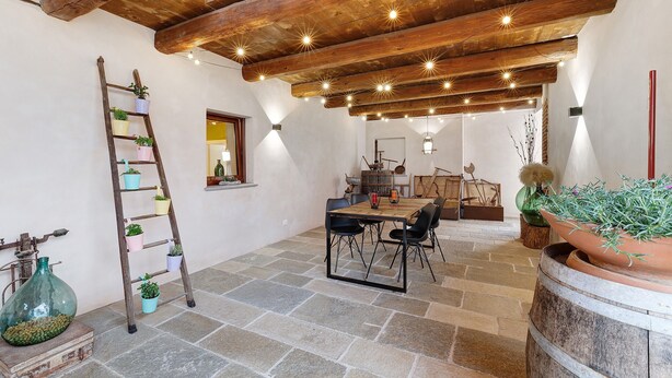 A rustic interior with exposed wooden beams, stone flooring, a wooden table with chairs, and potted plants.