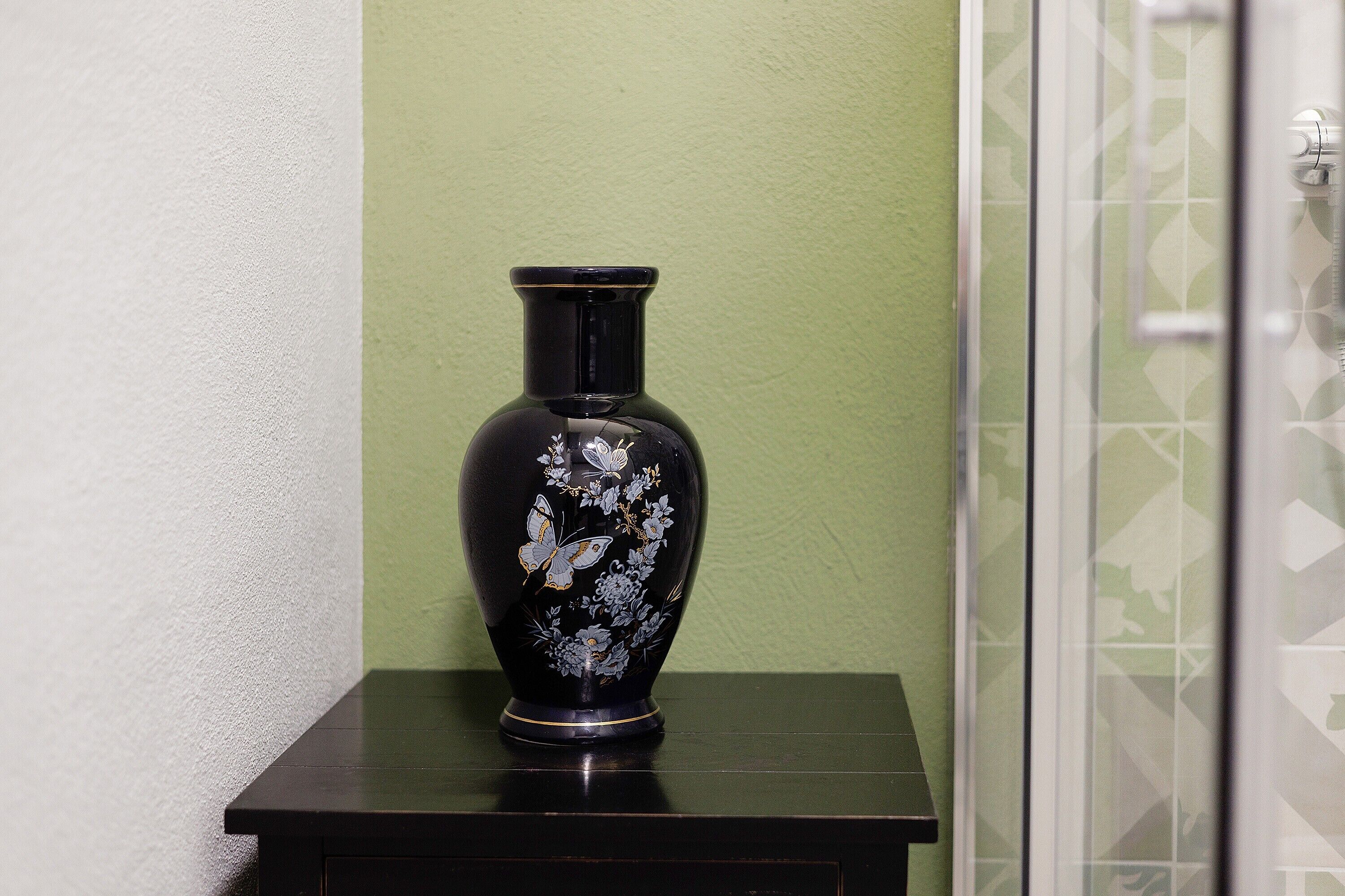 Un jarrón negro con diseños florales y de mariposas sobre una mesa de madera oscura frente a una pared verde.