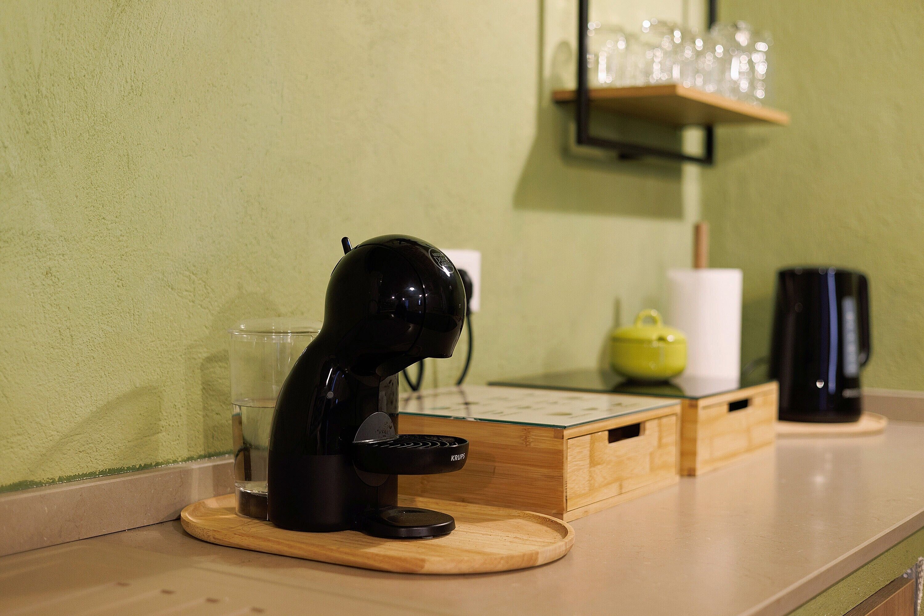 Une machine à café posée sur un plateau en bois, un verre, une théière jaune et une cafetière noire sur un plan de travail.