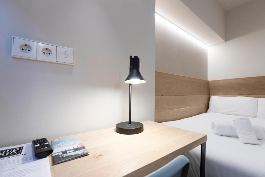 Habitación de hotel con cama, escritorio con lámpara, control remoto y folleto.