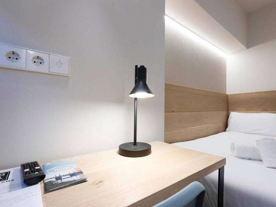 A hotel room with a bed, a desk with a lamp, a remote control, and a brochure.