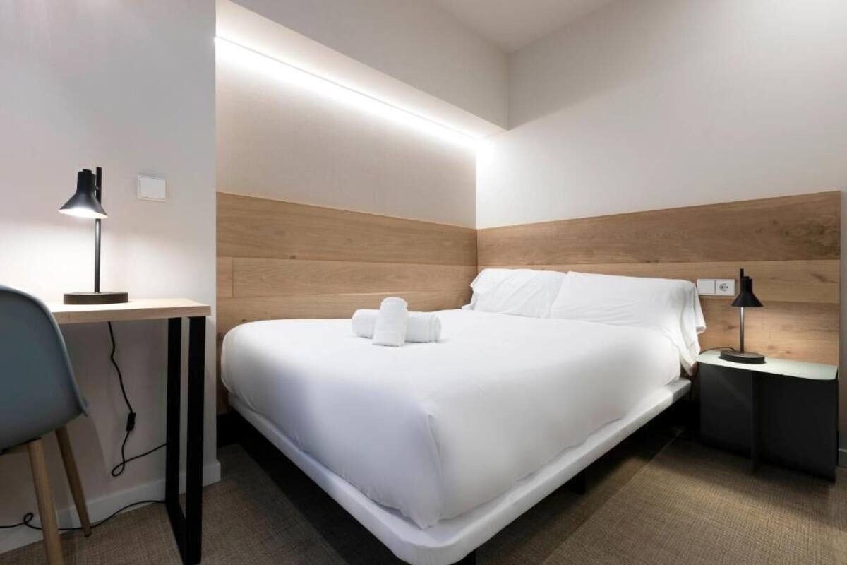Ein modernes Hotelzimmer mit einem ordentlich bezogenen Bett, einem Schreibtisch und einem Nachttisch mit Lampe.