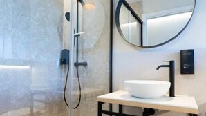 A modern bathroom with a glass shower enclosure, a white sink, a black faucet, a round mirror, and a red pendant light. - Urbaia Rooms (San Sebastian)