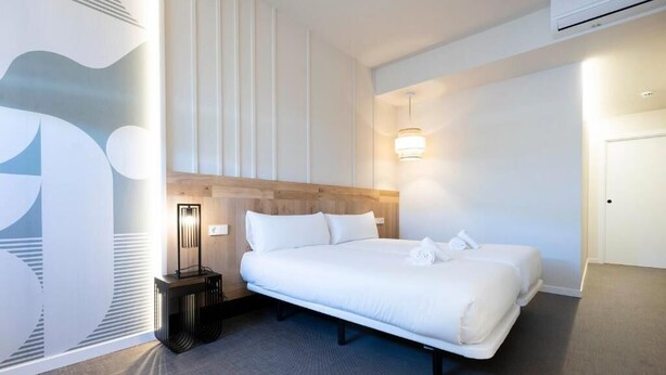A modern hotel room with a large bed, a bedside table, a wall-mounted light, and a door. - Urbaia Rooms (San Sebastian)