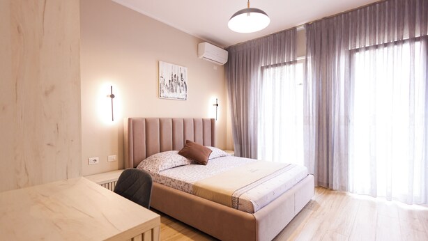 A modern bedroom with a bed, bedside lamps, a desk, and a large window with curtains. - Grandstay Urban Units (Durrës)