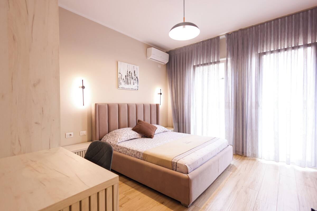 Um quarto moderno com cama, abajures, escrivaninha e uma janela grande com cortinas.