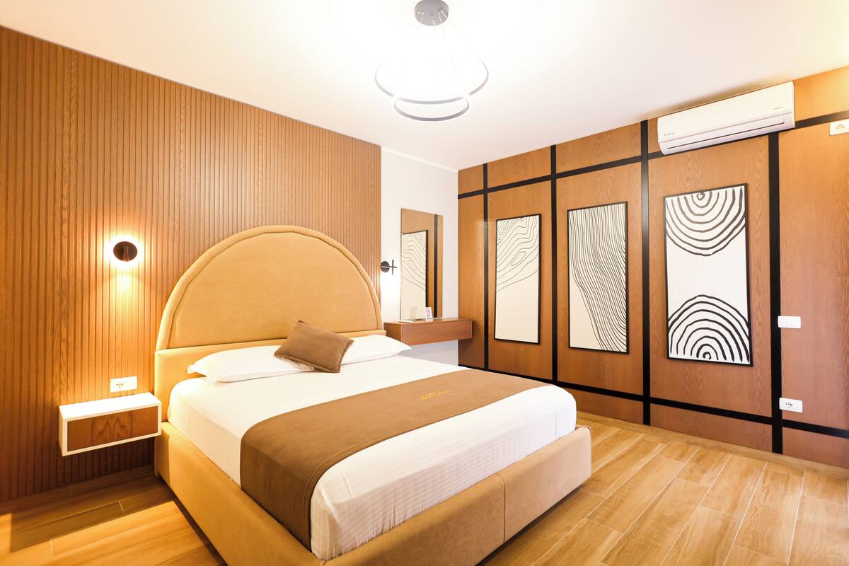 Quarto de hotel moderno com uma cama grande, painéis de madeira, mesa de cabeceira e obras de arte nas paredes.