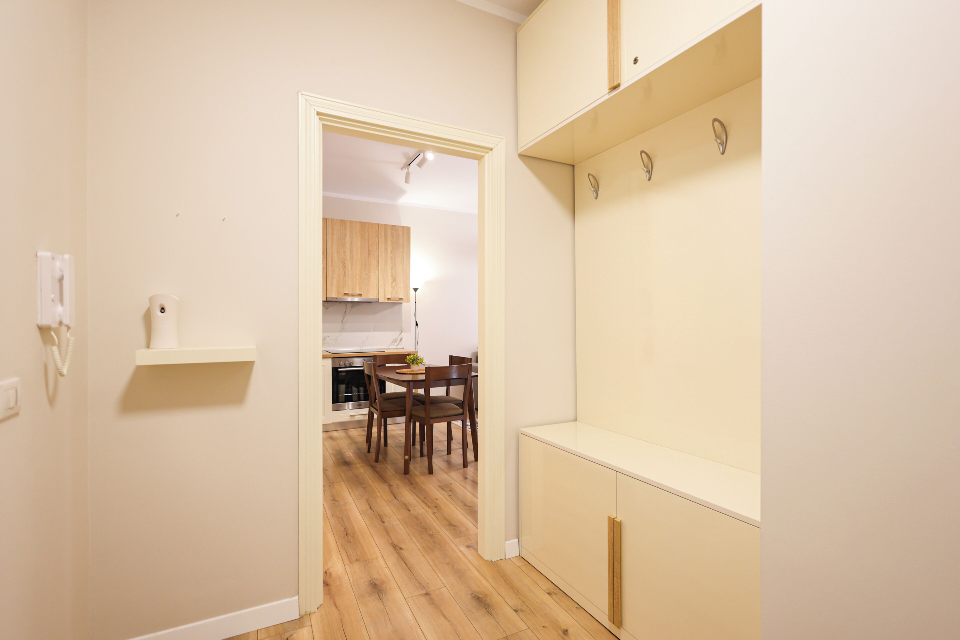 A narrow hallway with a wooden floor, a white wall, and a kitchen area visible through an open door.