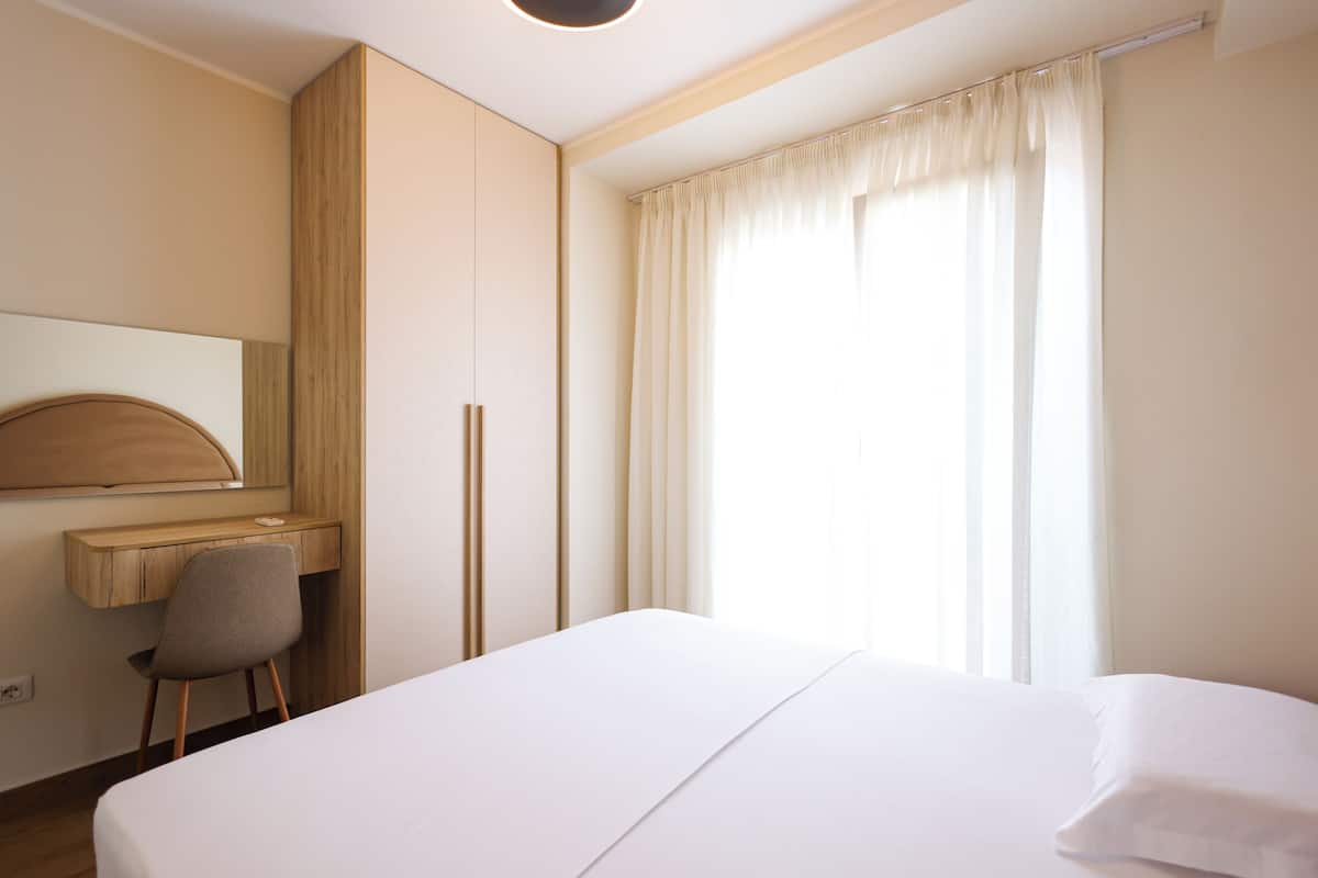 Quarto de hotel com cama, escrivaninha, cadeira, espelho e janela com cortinas.