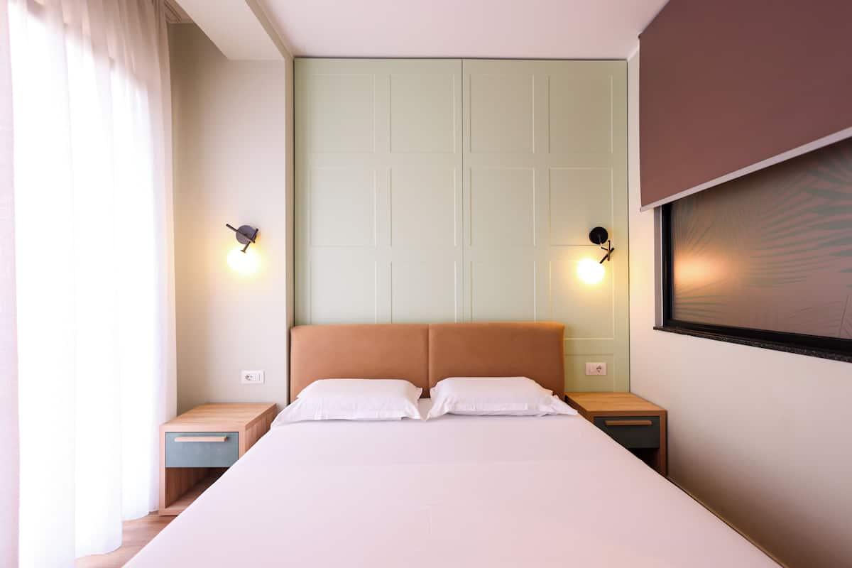 Quarto com cama, mesas de cabeceira, luminárias na parede e janela com cortinas.