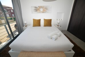 Ein Hotelzimmer mit einem großen Bett, zwei Kissen, einem Nachttisch mit Lampe, einem Wandbild und Blick auf Gebäude.