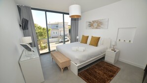 Une chambre moderne avec un grand lit, une table de chevet, une commode, un banc, une grande fenêtre et une vue sur l’extérieur.