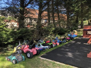 Eine Reihe von Kinderfahrzeugen steht auf einem befestigten Weg, flankiert von Grasflächen, umgeben von Bäumen und einem Gebäude im Hintergrund.