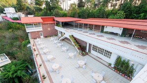 An outdoor patio with multiple round tables and chairs, surrounded by greenery and a building with a red roof. - DLS Tehri Club Resort (Tehri)