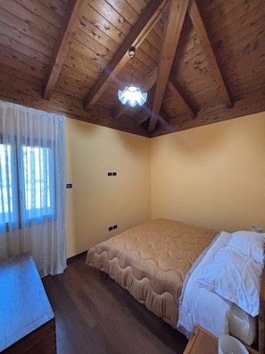 A bedroom with a wooden ceiling, a bed with a beige bedspread, a window with sheer curtains, and a bedside table.