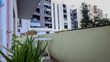 Un balcón con una planta en maceta, una mesa blanca y vistas a un edificio moderno con varios balcones.
