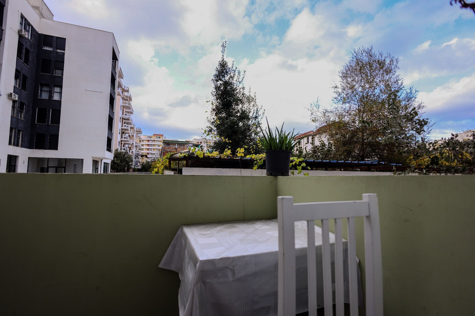 Un balcón con una mesa y silla blancos, con vista a un paisaje urbano con edificios y árboles.