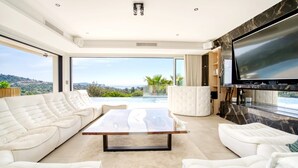 A modern living room with a large TV, white sofas, a glass coffee table, and a view of the outdoors. - Villa Les Oiseaux Du Paradis Cavalaire-sur-mer (Cavalaire-sur-Mer)