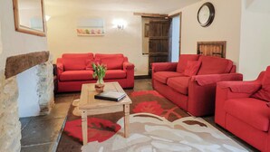 A living room with two red sofas, a wooden coffee table, a vase with flowers, a clock on the wall, and a rustic wooden door. - Cider Cottage - Near Lyme Regis (Axminster)