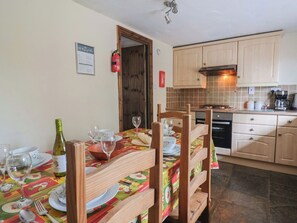 A dining area with a table set for four, wooden chairs, a bottle of wine, and glasses. A kitchen with light wooden cabinets and a stove is visible in the background. - Cider Cottage - Near Lyme Regis (Axminster)