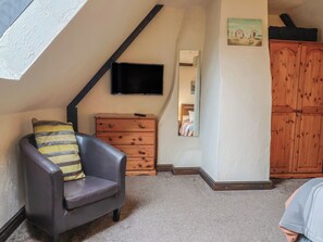 A room with a sloped ceiling, a television mounted on the wall, a wooden dresser, a chair with a cushion, and a wardrobe. - Cider Cottage - Near Lyme Regis (Axminster)