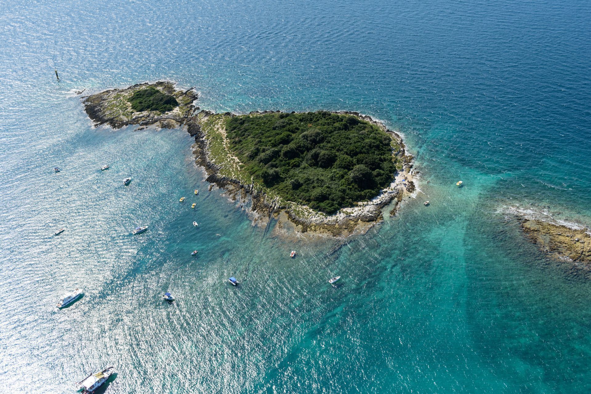 Luftaufnahme einer Küstenlandschaft mit klarem türkisblauem Wasser, Felsformationen und einer kleinen Insel mit dichter Vegetation.