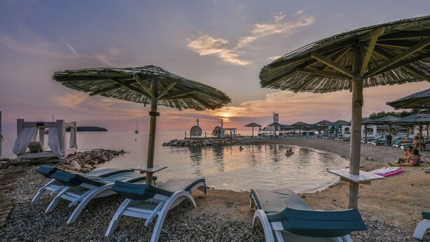 Uma praia com cadeiras de praia e guarda-sóis, um mar calmo e um pôr do sol ao fundo.