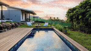 Una piscina con cubierta de madera, dos sillones de descanso y una casa moderna al fondo.