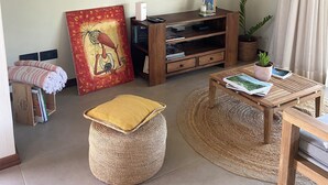 Un salón con televisión de pantalla plana, mueble de madera para equipo multimedia, un cuadro grande, una planta en maceta y un reposapiés tejido.