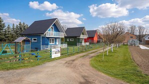 Maalaismaisema, jossa on hiekkapolku, joka johtaa värikkäisiin taloihin, aidattu leikkikenttä ja kirkas taivas.