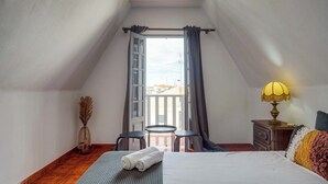 A bedroom with a bed, a small table, a lamp, a vase, and a view of the outside.