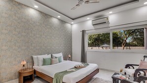 A modern bedroom with a large bed, bedside tables, a window with a view, and an air conditioning unit. - Alaya Stays Ratikara Ullaas 2BHK Pool (Ballabgarh)