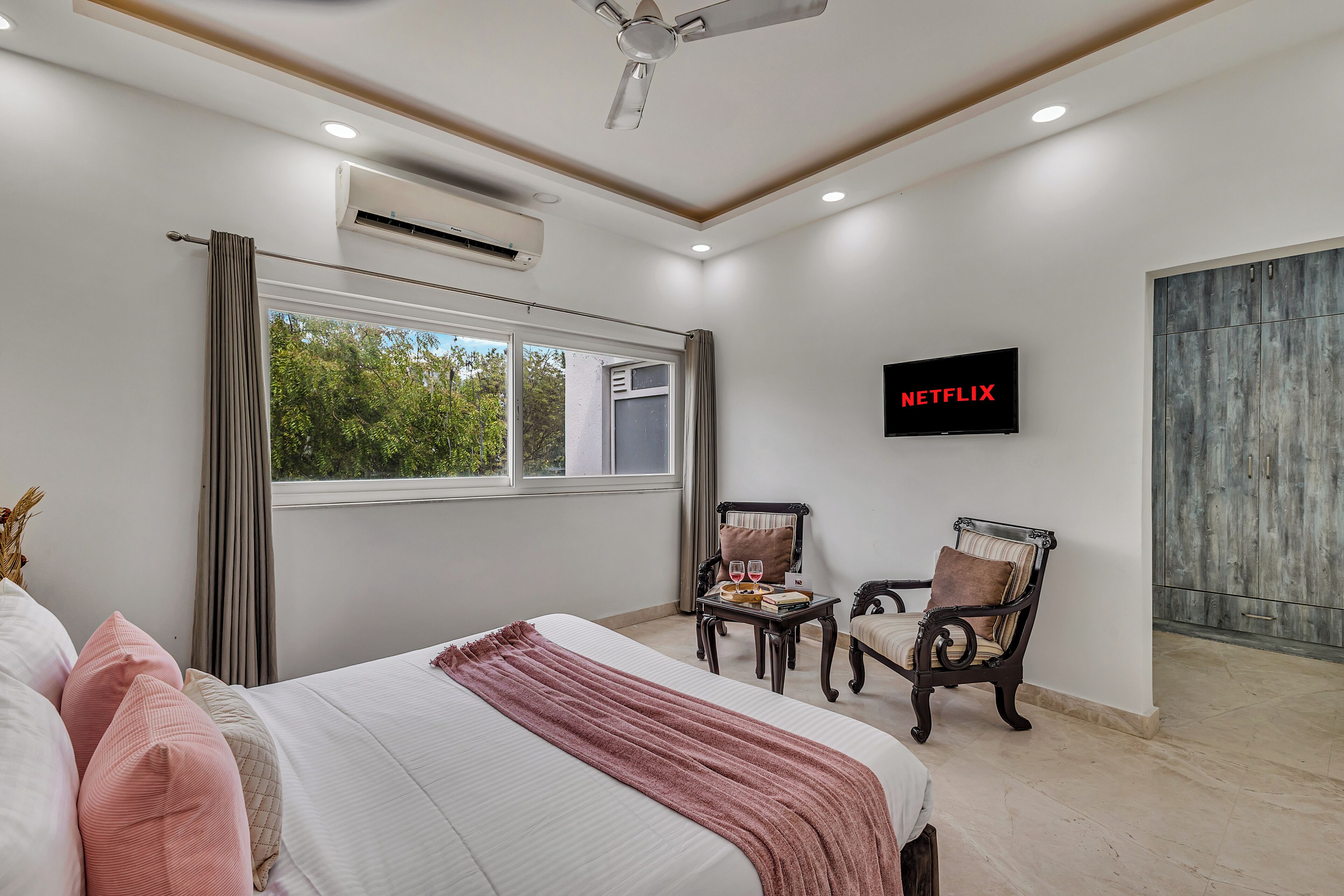 A hotel room with a bed, a TV, a window, and a view of greenery outside.