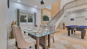 A dining area with a glass table, cushioned chairs, a billiard table, and a staircase with a decorative railing. - Alaya Stays Ratikara Ullaas 2BHK Pool (Ballabgarh)