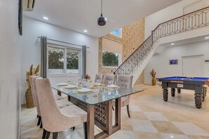 A dining area with a glass table, cushioned chairs, a billiard table, and a staircase with a decorative railing.