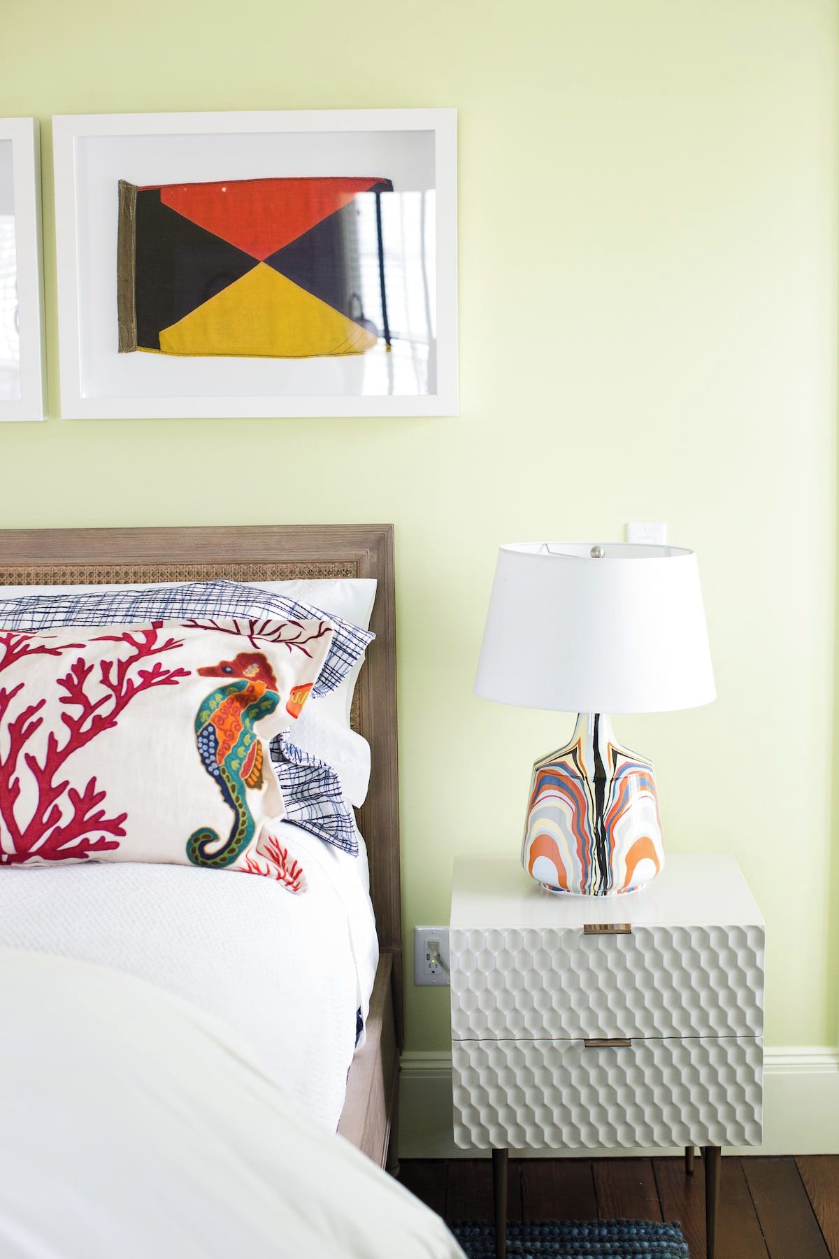 A neatly made bed with a colorful seahorse pillow, a white lamp with a patterned base, and a geometric artwork on the wall.
