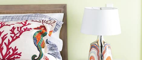 A neatly made bed with a colorful seahorse pillow, a white lamp with a patterned base, and a geometric artwork on the wall.
