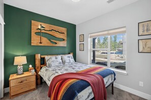 Un dormitorio con una cama, una mesita de noche de madera, una ventana con vistas y una obra de arte colgada en la pared.