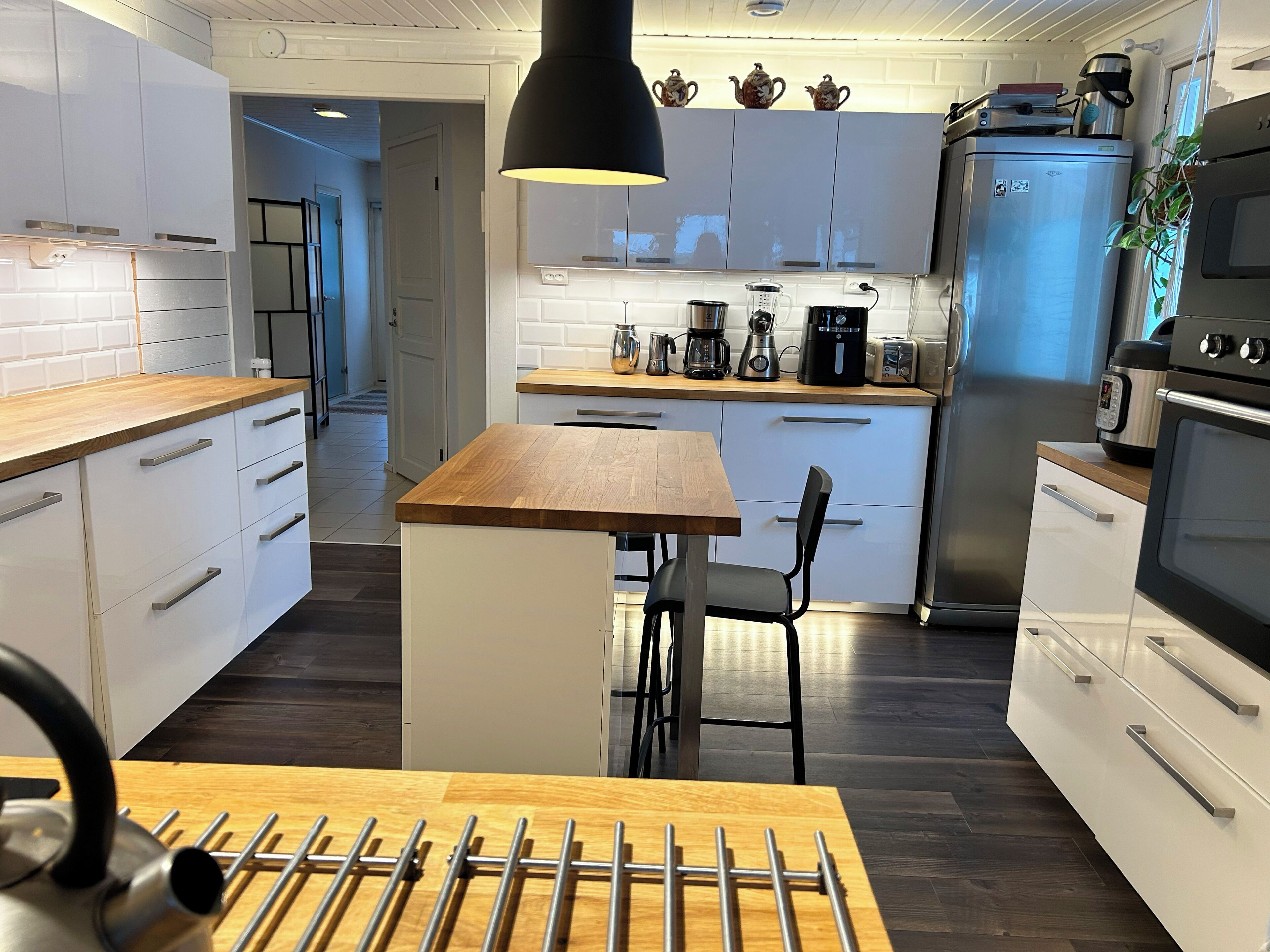Una cocina moderna con armarios blancos, una isla de madera, electrodomésticos de acero inoxidable y una lámpara colgante negra.