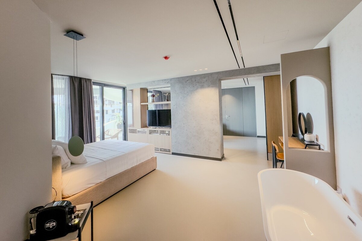 A modern hotel room with a large bed, a sitting area, a kitchen, and a bathroom.