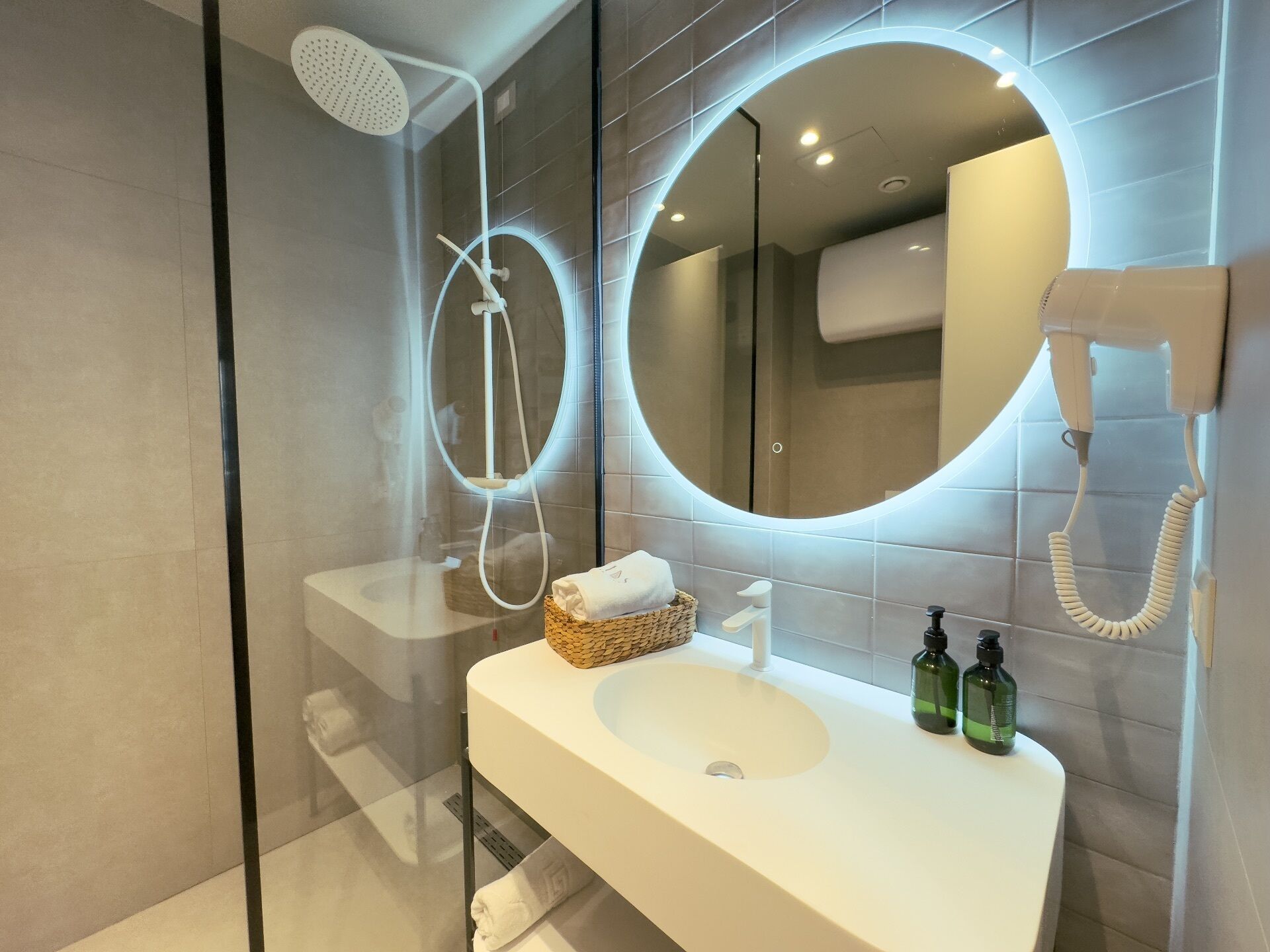 A modern bathroom with a glass-enclosed shower, a round mirror, a hairdryer, and a sink with toiletries.