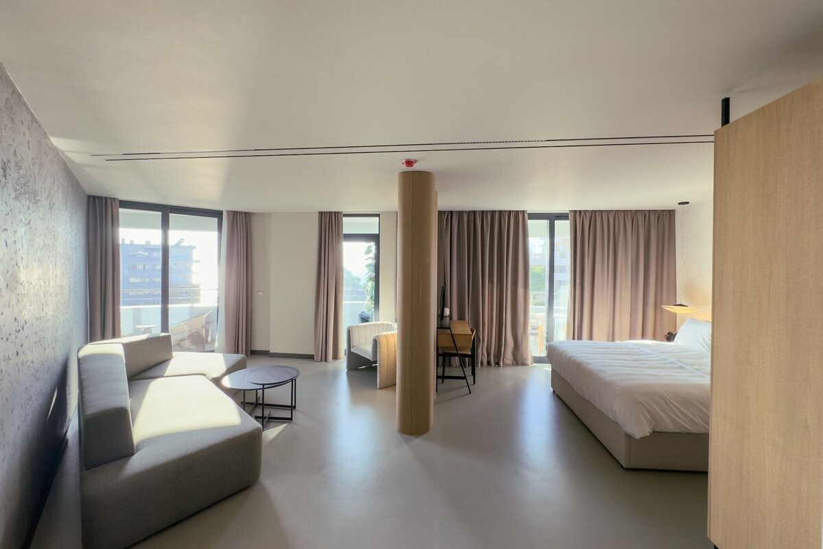 A hotel room with a bed, a sofa, a small table, and a chair. There is a large window with curtains, and the room has a modern design.