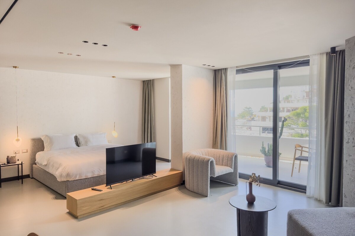 A modern hotel room with a bed, a TV, a seating area, and a balcony with a view.