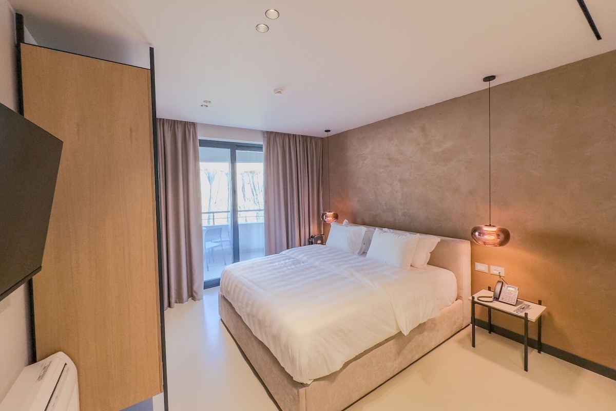A modern hotel room with a large bed, a desk with a telephone, and a view of the outdoors.