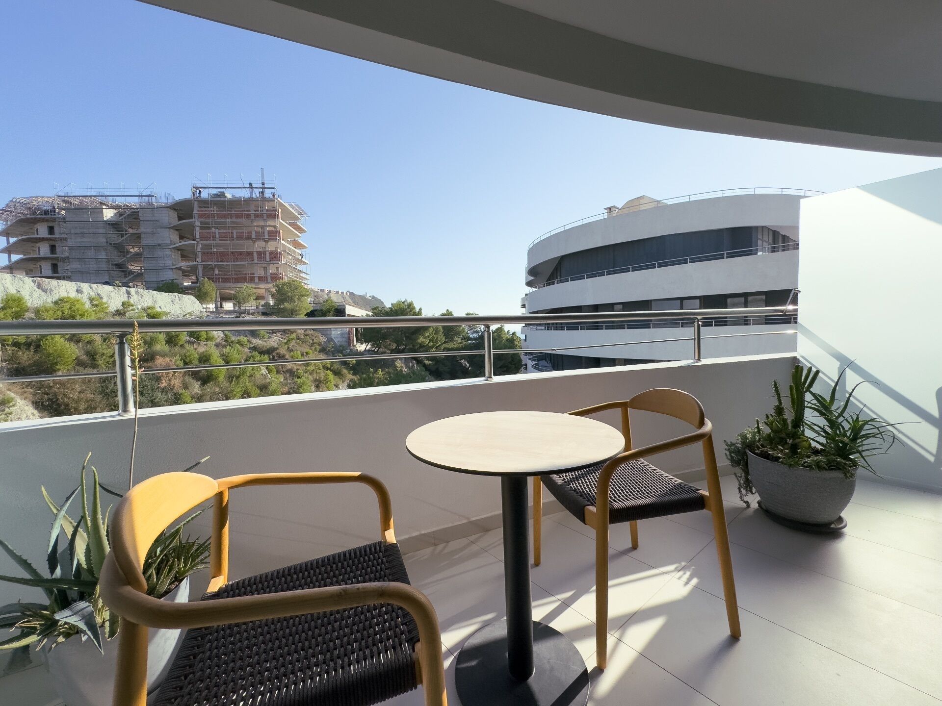 A balcony with two chairs and a table, overlooking a building under construction and a modern building.