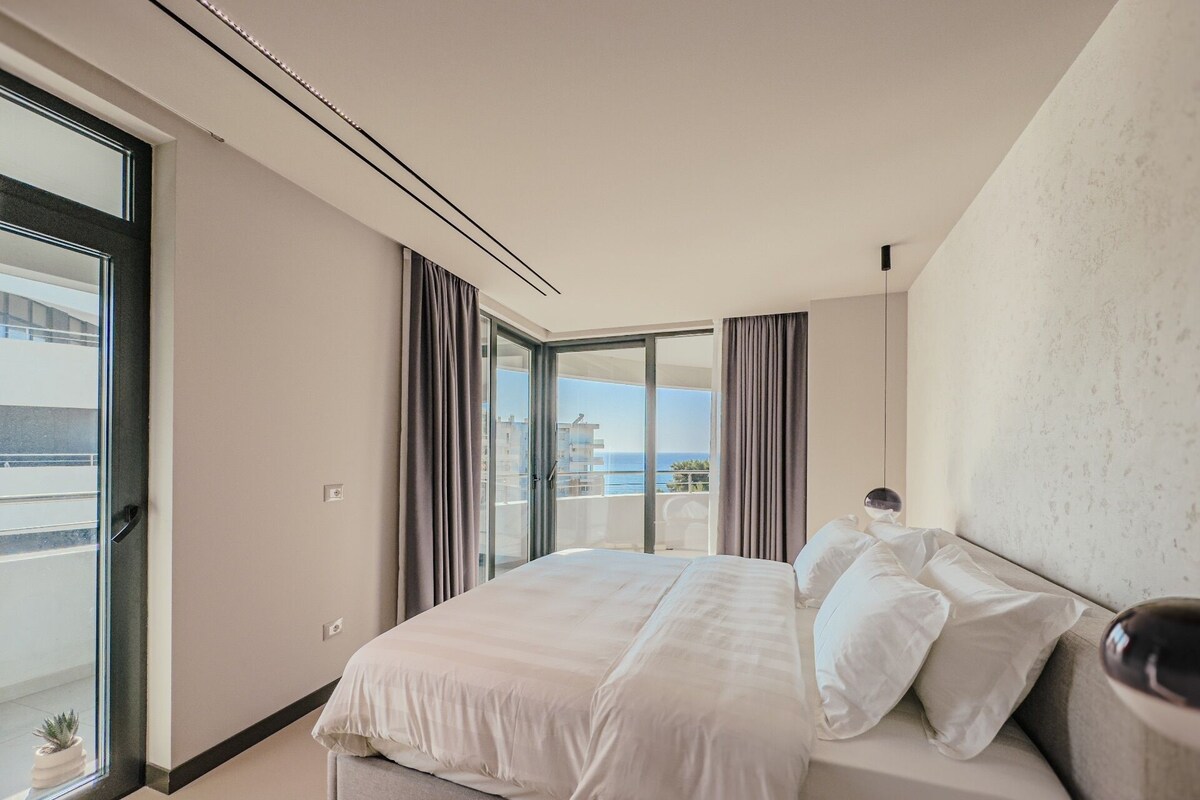 A modern hotel room with a large bed, a view of the sea, and a balcony.