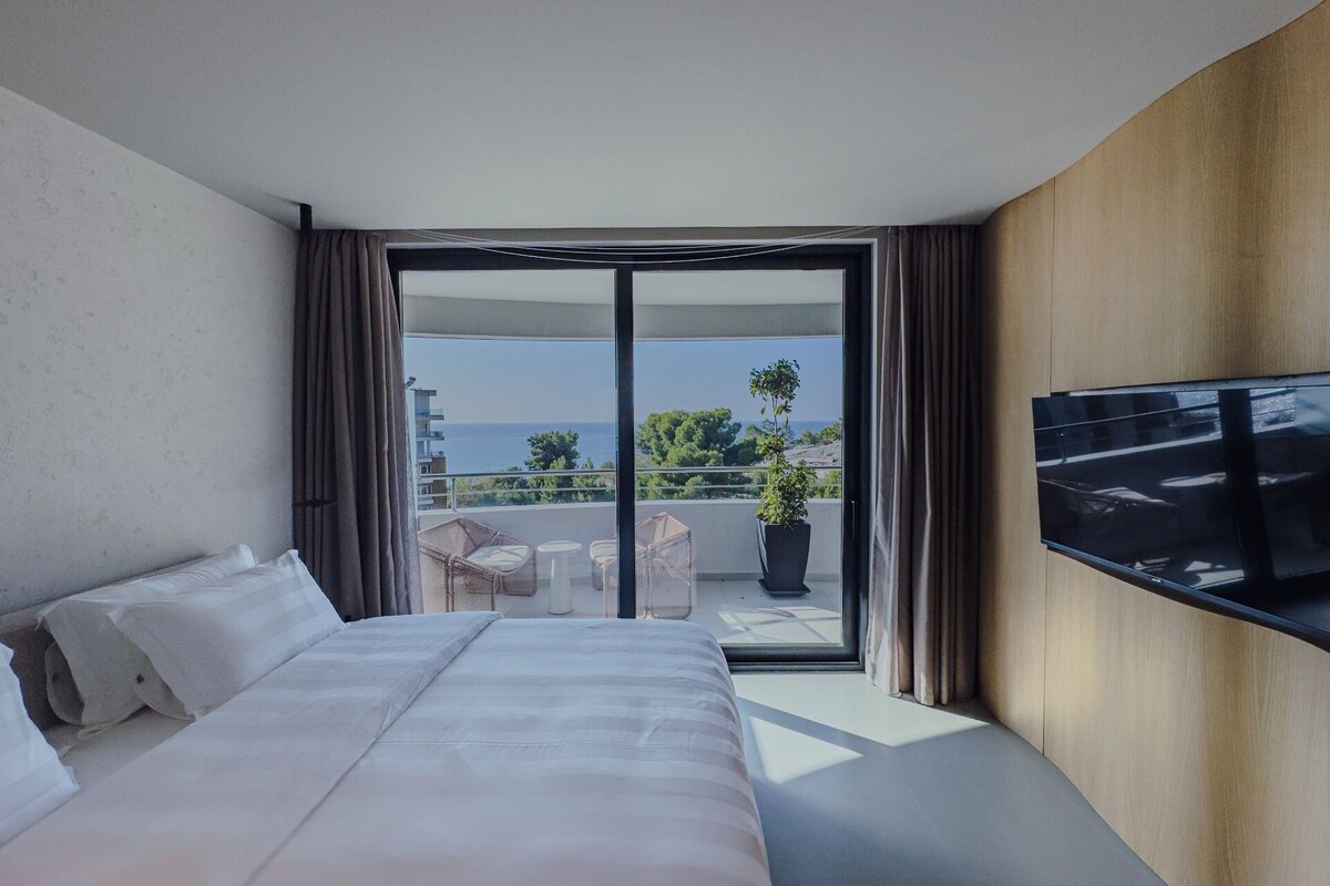 A hotel room with a large bed, a TV mounted on the wall, and a balcony with a view of the sea.