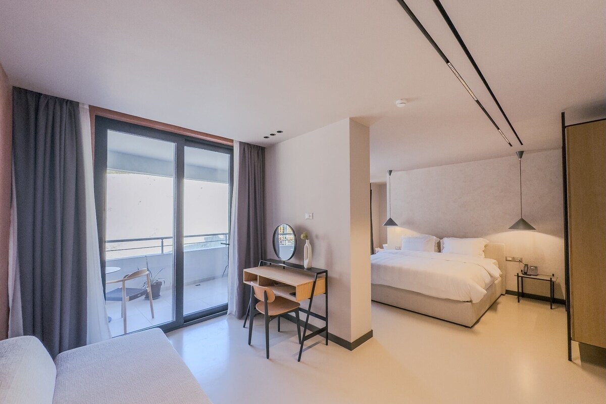 A modern hotel room with a balcony, a desk with a chair, a bed, and a bedside table.