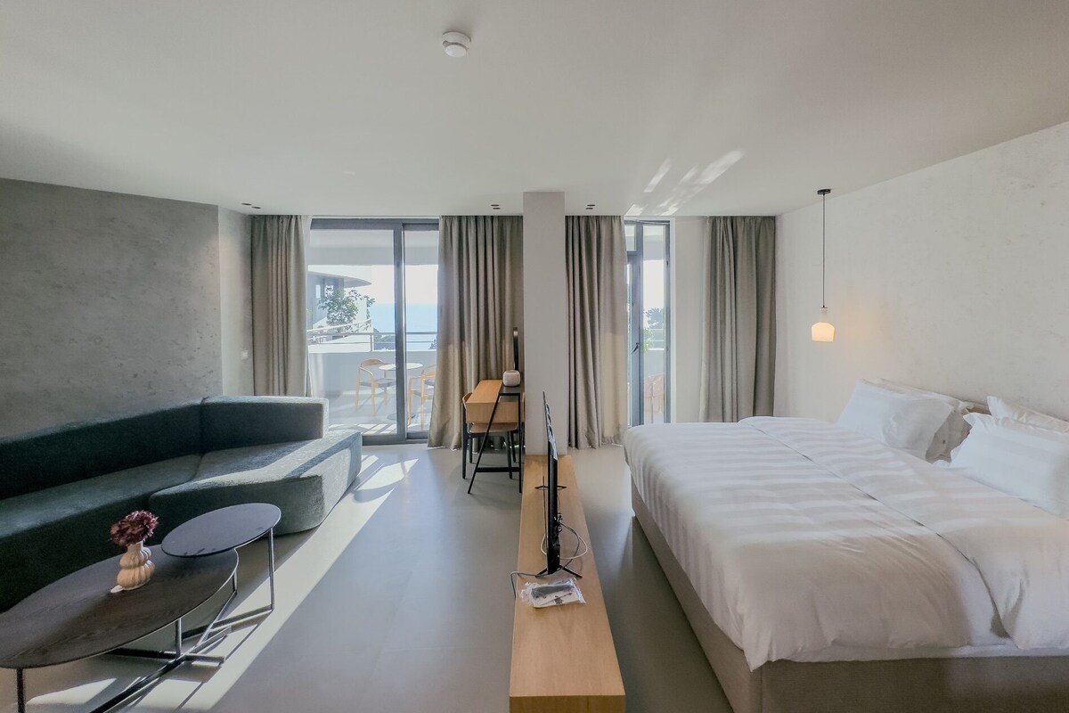 A modern hotel room with a bed, a sofa, a desk, and a view of the outdoors.
